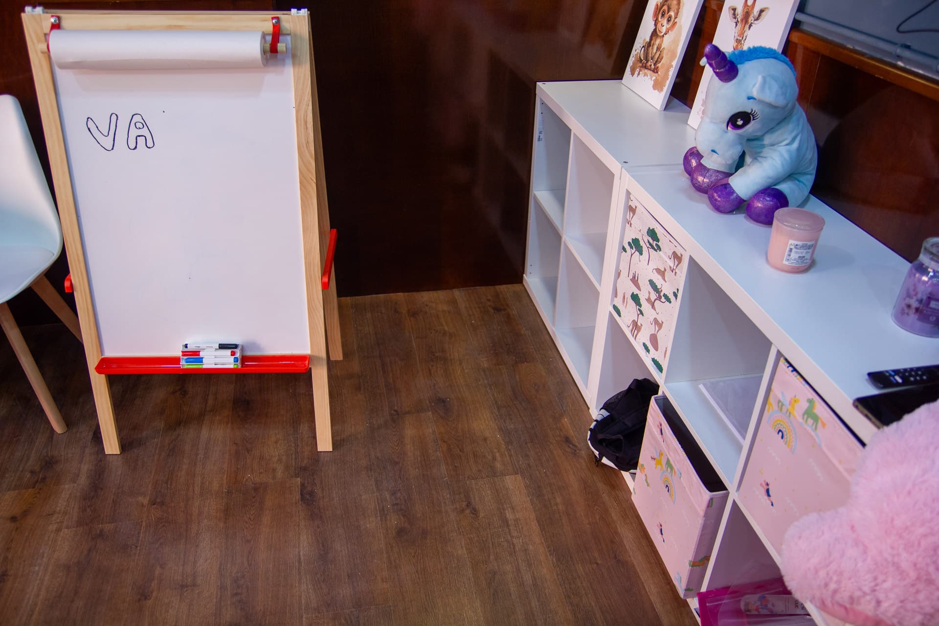 Angolo gioco fuori dalla grotta: cavalletto con lavagna bianca e scaffale bianco con peluche, scatole decorate e disegni di animali.
