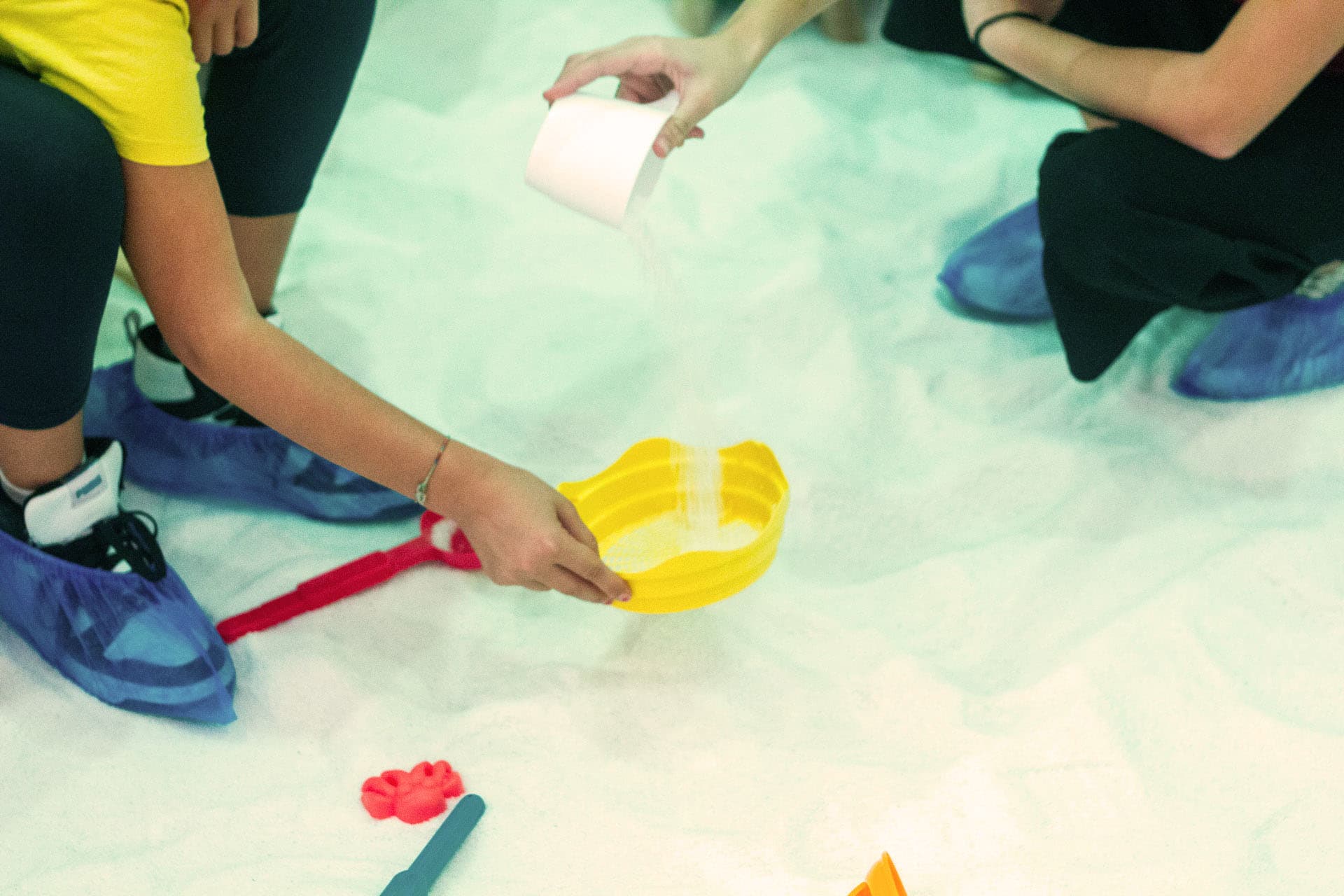 Primo piano di mani che giocano sul sale: un adulto versa sale in un secchiello giallo tenuto da un bambino; sul pavimento palette e formine colorate.