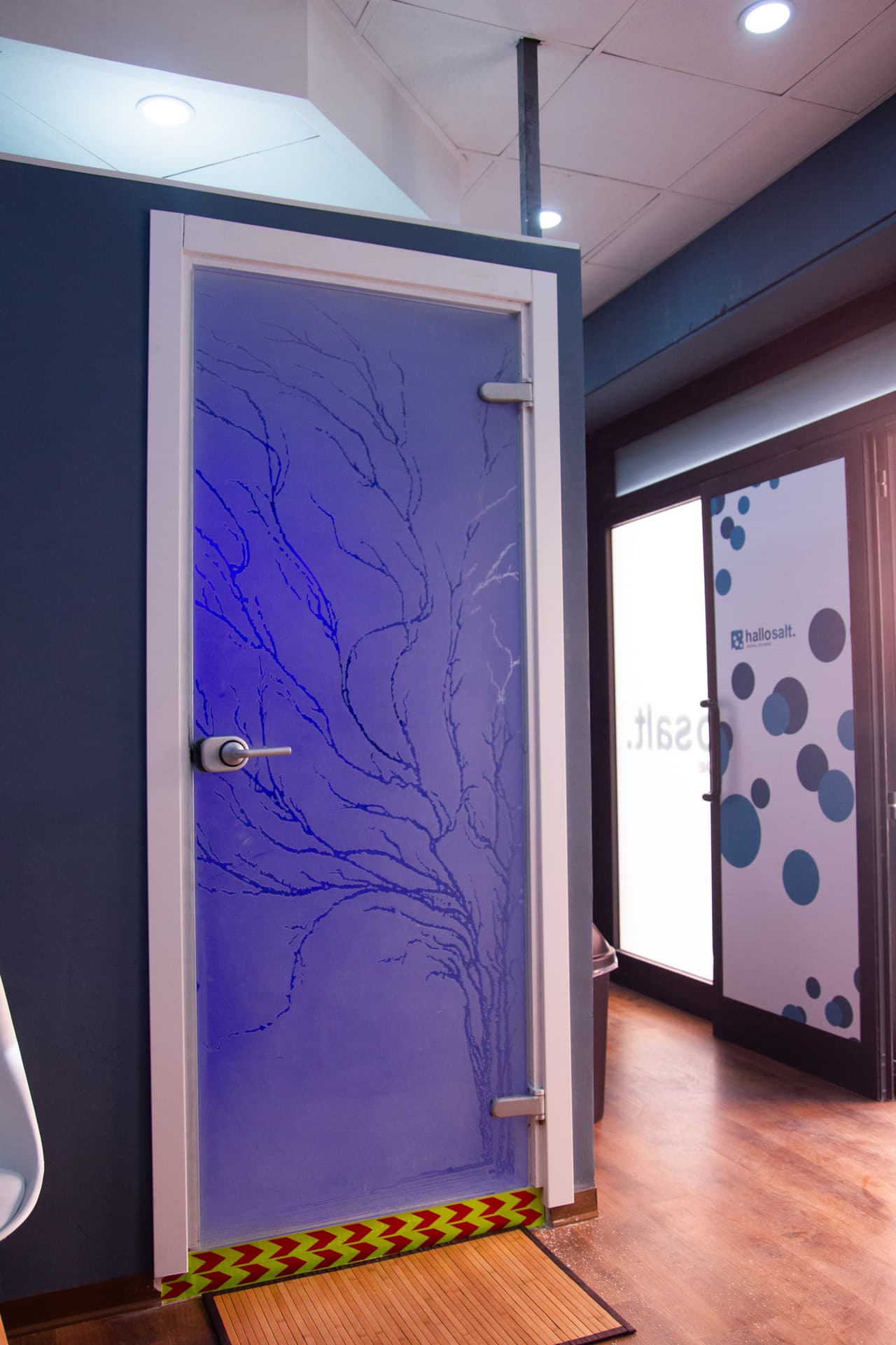 Porta a vetro satinato con motivo ad albero e luce blu-viola dall’interno; a destra, vetrata con logo hallosalt. e pois blu.