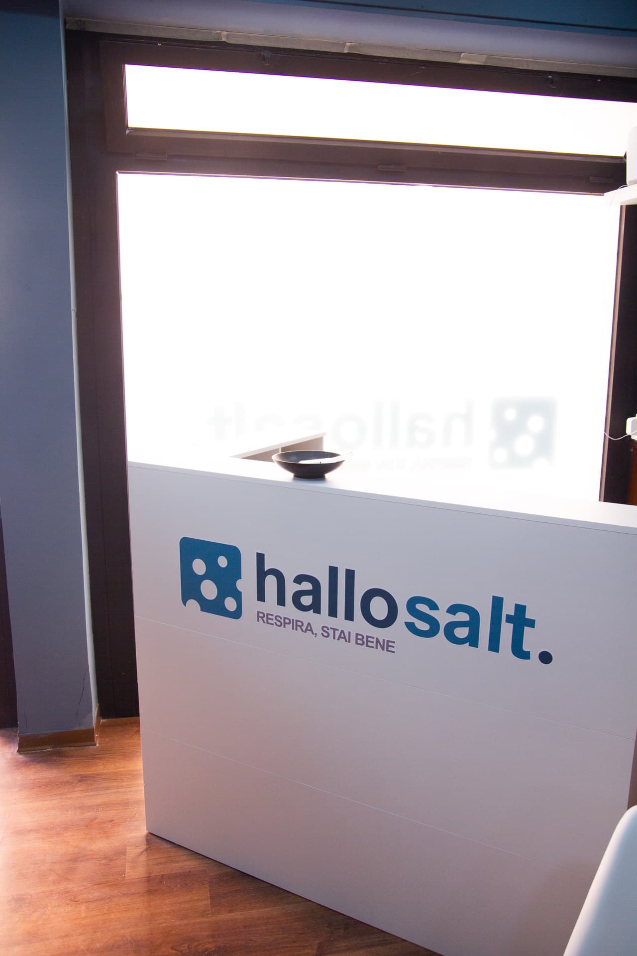 Bancone bianco della reception Hallosalt con logo e motto «Respira, stai bene», davanti a una vetrata smerigliata.