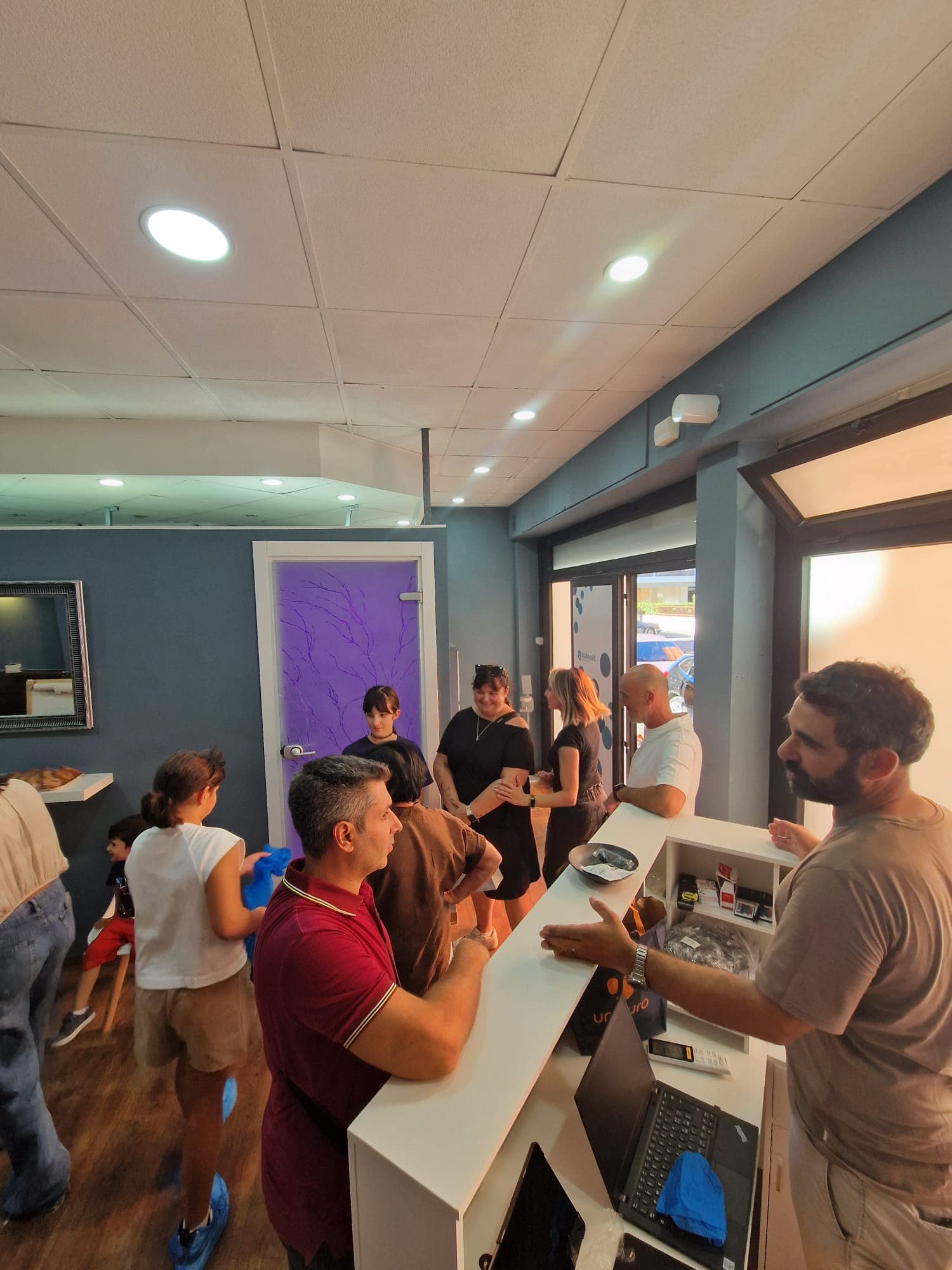 Persone riunite alla reception Hallosalt: bancone bianco, pareti blu e porta viola con motivo ad albero sullo sfondo.
