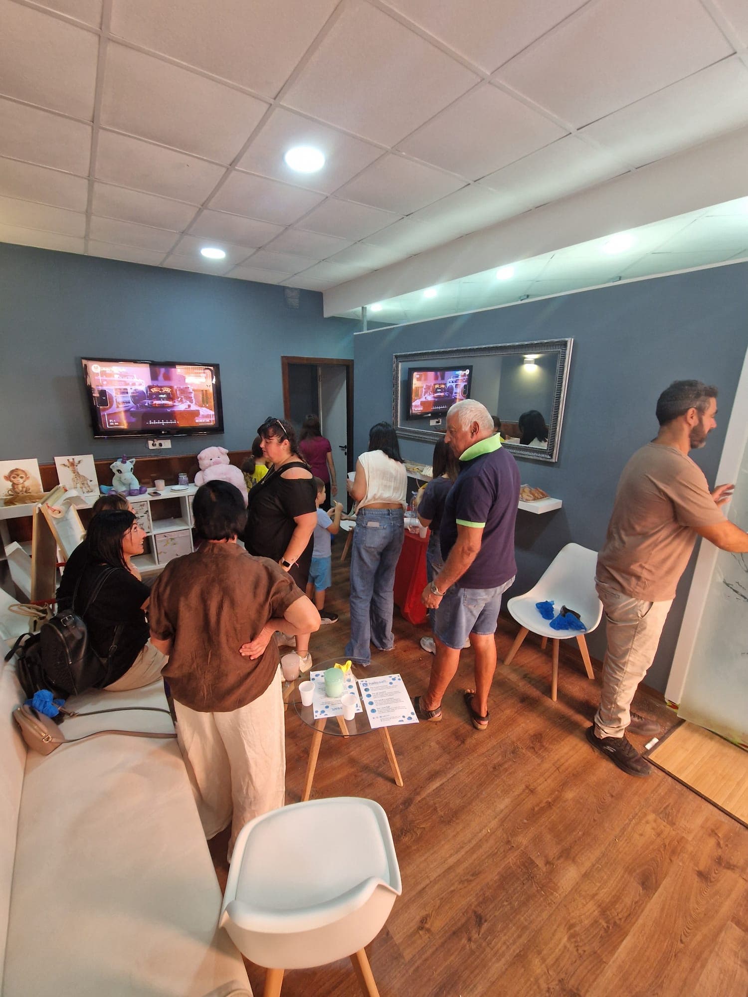 Sala d’attesa con pareti blu e pavimento in legno: adulti e bambini in conversazione, panca bianca, TV e scaffale con peluche.
